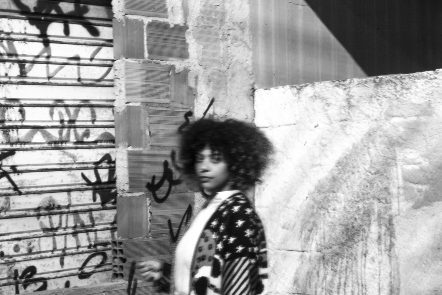 Fotografia em preto e branco da artista Thati Dias. Ela é uma mulher negra, de cabelos crespos encaracolados e veste blusa estampada. Fotografia em preto e branco da artista Thati Dias. Ela é uma mulher negra, de cabelos crespos encaracolados e veste blusa estampada.