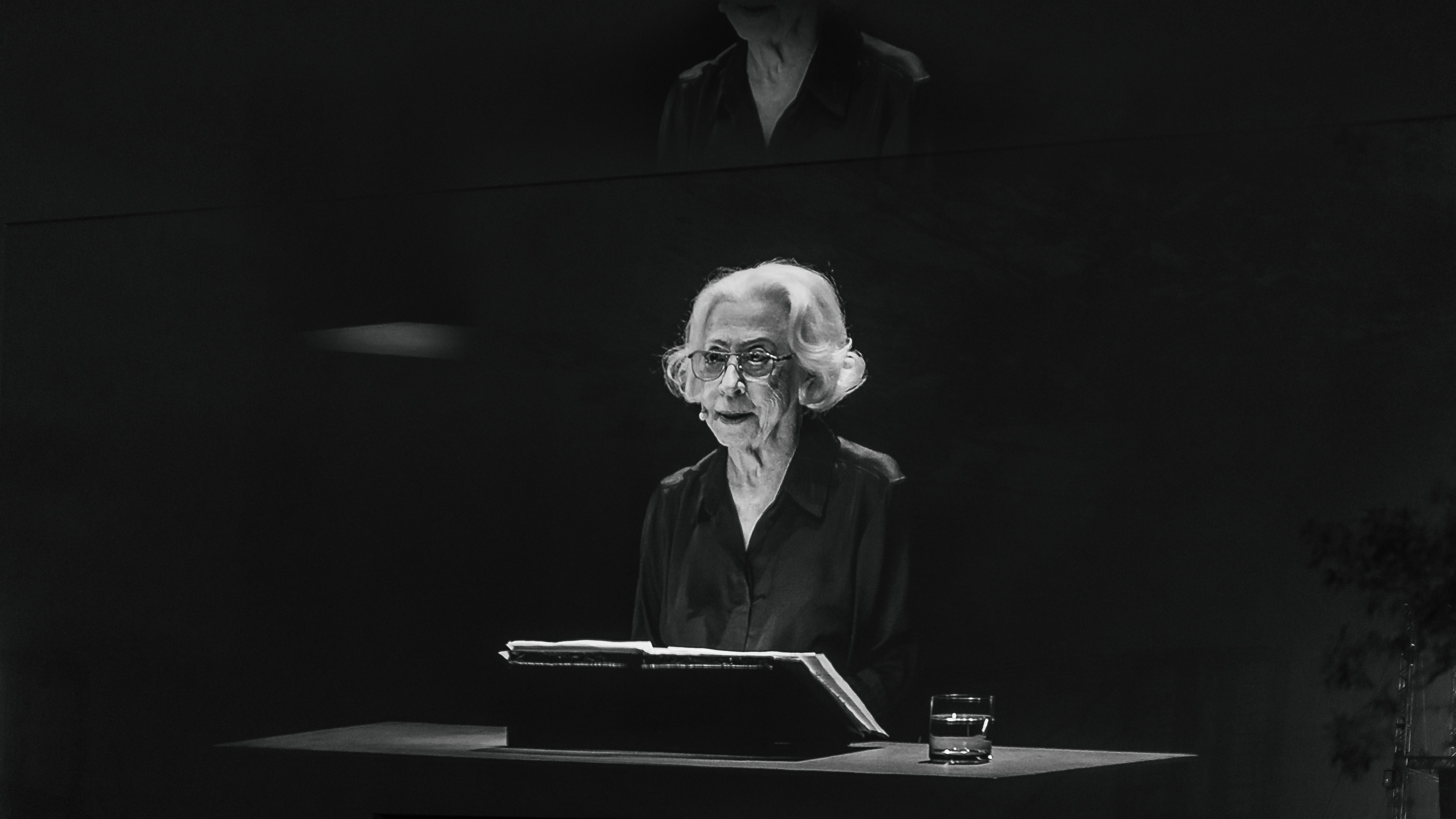Foto preto e branca mostra Fernanda Montenegro, mulher idosa de cabelos brancos e lisos na altura do queixo. Ela usa óculos de grau e camisa preta. Está sentada diante de uma mesa preta. Há um livro aberto à sua frente, assim como um copo com água do seu lado direito. Foto preto e branca mostra Fernanda Montenegro, mulher idosa de cabelos brancos e lisos na altura do queixo. Ela usa óculos de grau e camisa preta. Está sentada diante de uma mesa preta. Há um livro aberto à sua frente, assim como um copo com água do seu lado direito.