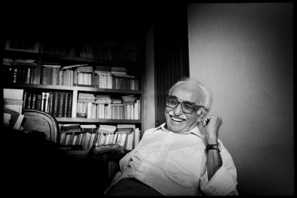 Manoel de Barros aparece sentado, sorrindo, vestindo uma camisa e usando óculos e relógio. Atrás dele, há uma estante cheia de livros. Manoel de Barros aparece sentado, sorrindo, vestindo uma camisa e usando óculos e relógio. Atrás dele, há uma estante cheia de livros.