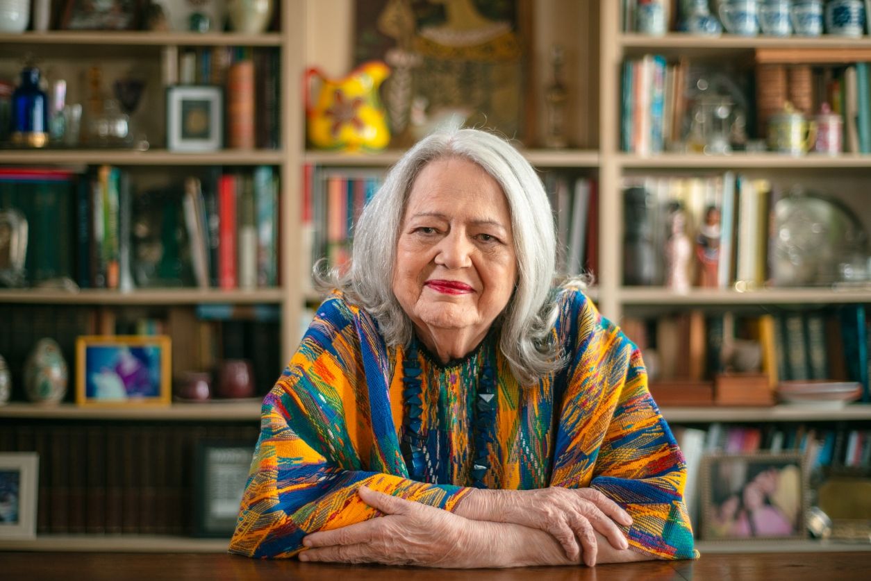 Fotografia colorida de Ana Mae Barbosa. Ela é uma mulher branca, idosa, possui cabelos brancos. Está usando uma roupa colorida, com tons de laranja, azul e verde, e um colar azul.