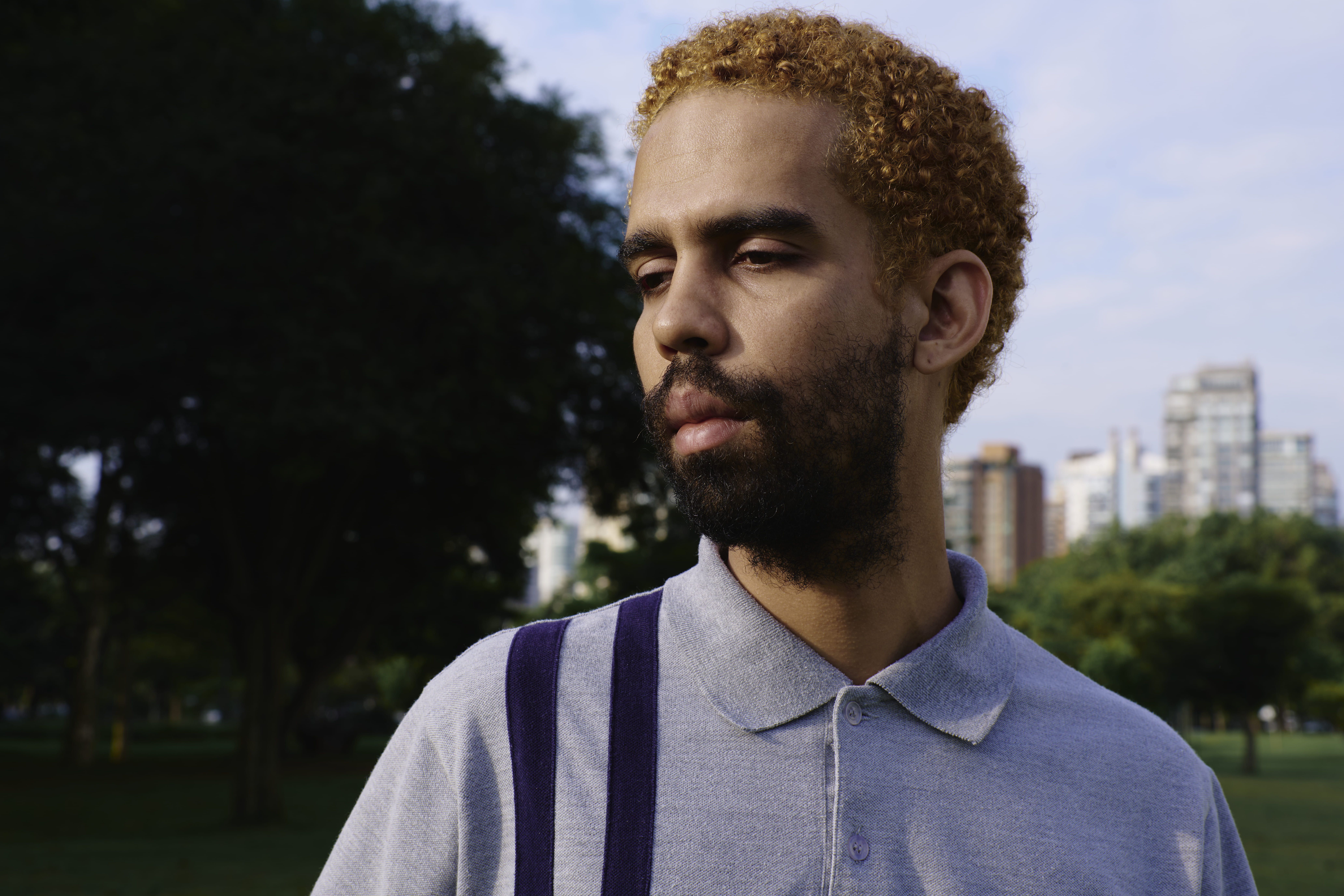 Homem jovem e negro com a pele clara aparece em foto dos ombros para cima, em parque. Ele usa camiseta pólo cinza, com duas listras azuis verticais. Ele tem barba escura e os cabelos crespos e curtos pintados de louro escuro. Homem jovem e negro com a pele clara aparece em foto dos ombros para cima, em parque. Ele usa camiseta pólo cinza, com duas listras azuis verticais. Ele tem barba escura e os cabelos crespos e curtos pintados de louro escuro.
