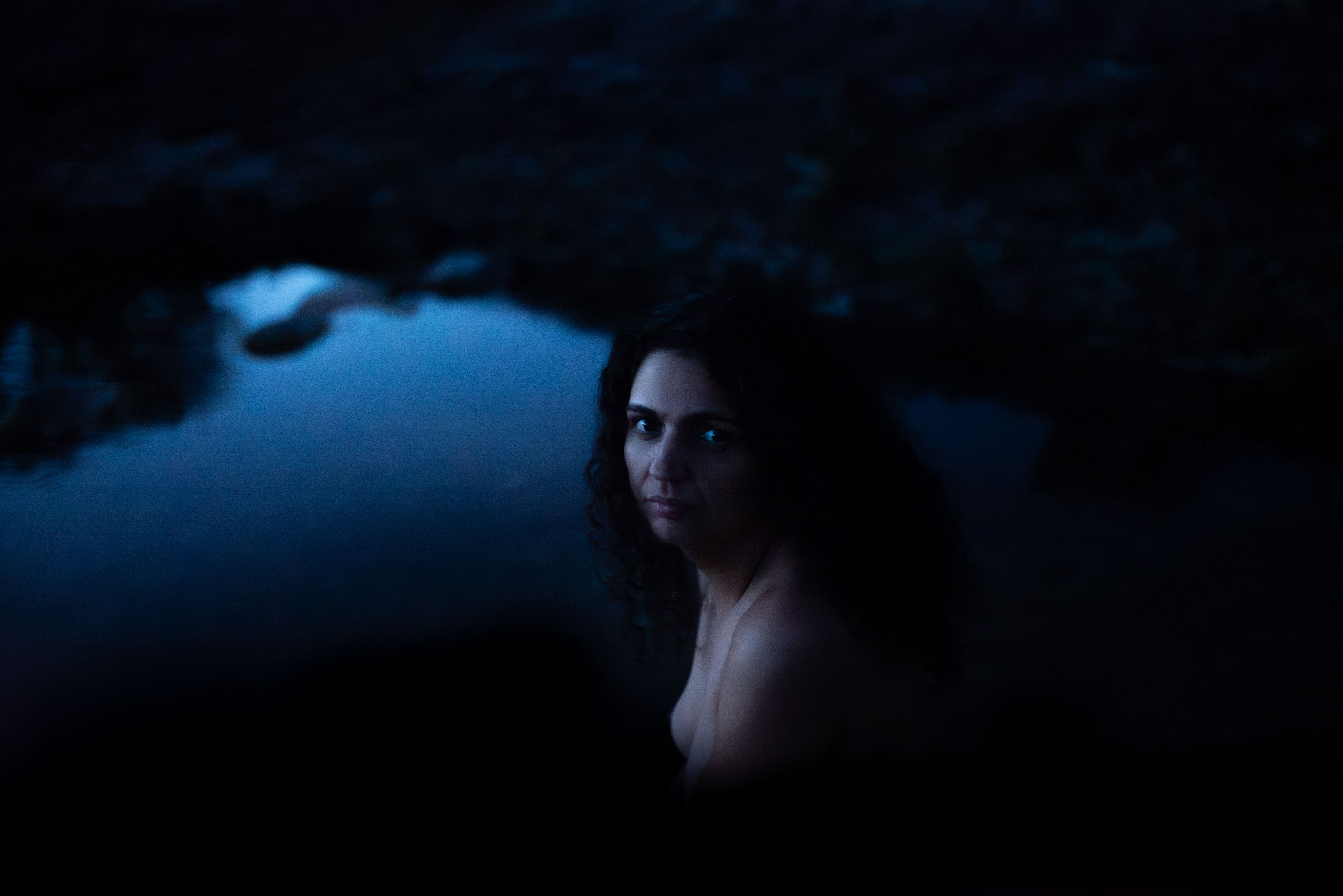 Luiza Brina aparece perto de águas. Ela está nua e olha séria para a câmera. A artista tem cabelo encaracolado. A imagem trabalha com sombras e falta de luz. Luiza Brina aparece perto de águas. Ela está nua e olha séria para a câmera. A artista tem cabelo encaracolado. A imagem trabalha com sombras e falta de luz.