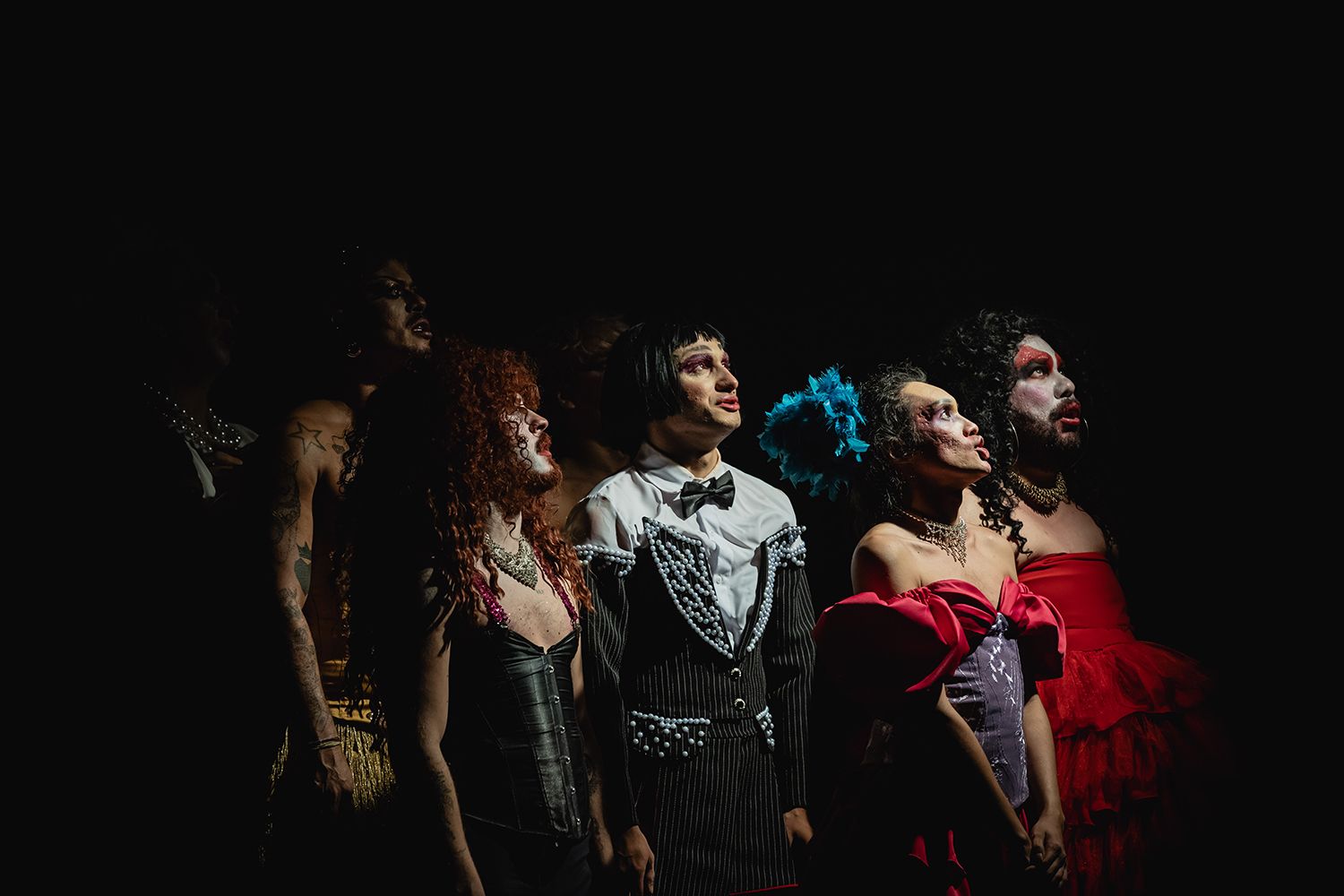 Fotografia colorida de cena do espetáculo Sebastião. Na imagem, sete pessoas vestida como drag estão com o corpo virado para a direita e olha para cima. Fotografia colorida de cena do espetáculo Sebastião. Na imagem, sete pessoas vestida como drag estão com o corpo virado para a direita e olha para cima.