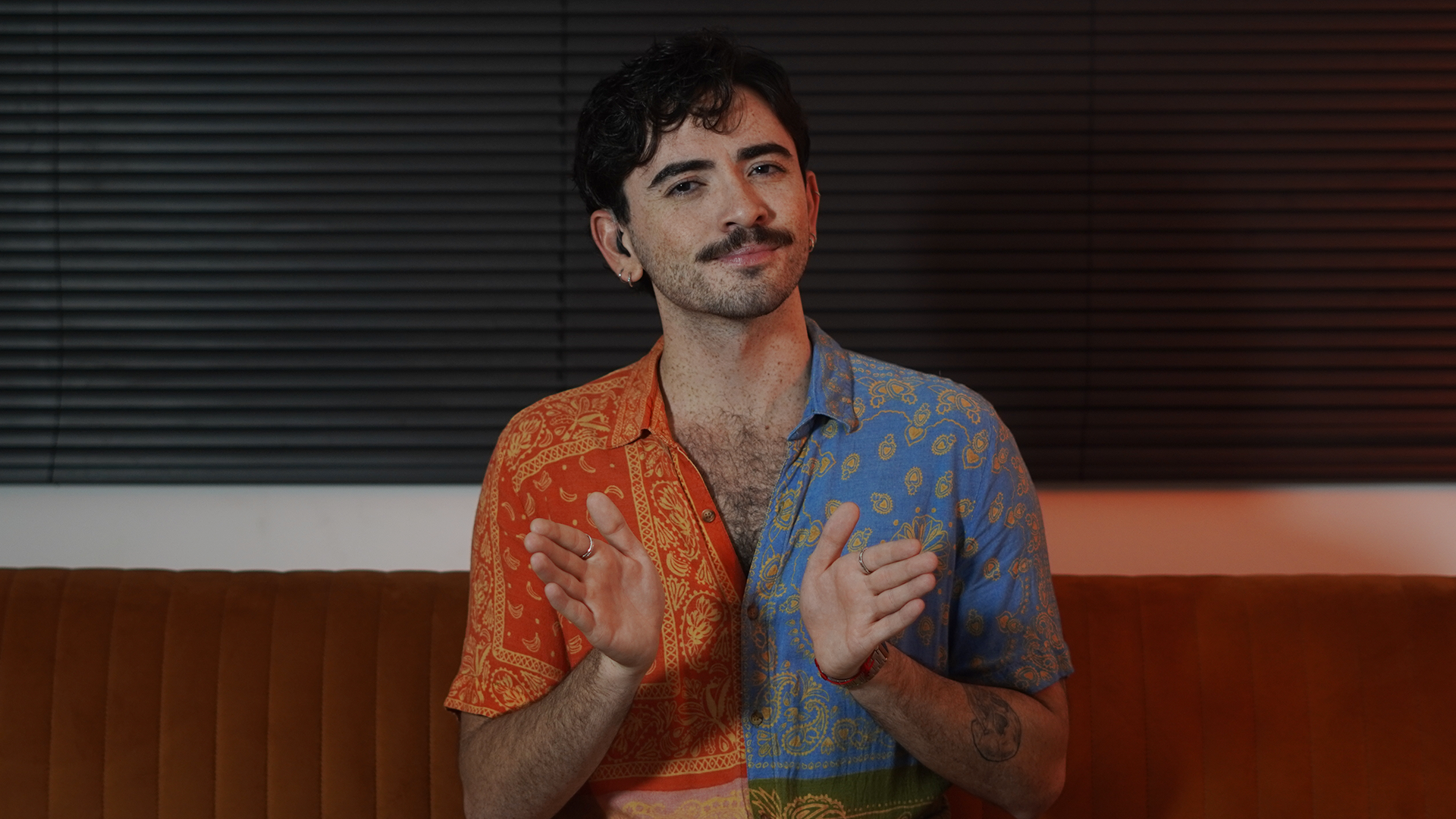 Homem jovem e branco aparece da cintura para cima, aparentemente sentado em um sofá de couro marrom. Ele tem cabelos curtos e pretos e usa bigode, também preto. Atrás dele, acima do sofá, tem uma persiana preta. Ele usa uma camisa de botão estampada e colorida. Tem uma tatuagem no braço e está sinalizando em Libras, gesticulando com as duas mãos. Homem jovem e branco aparece da cintura para cima, aparentemente sentado em um sofá de couro marrom. Ele tem cabelos curtos e pretos e usa bigode, também preto. Atrás dele, acima do sofá, tem uma persiana preta. Ele usa uma camisa de botão estampada e colorida. Tem uma tatuagem no braço e está sinalizando em Libras, gesticulando com as duas mãos.